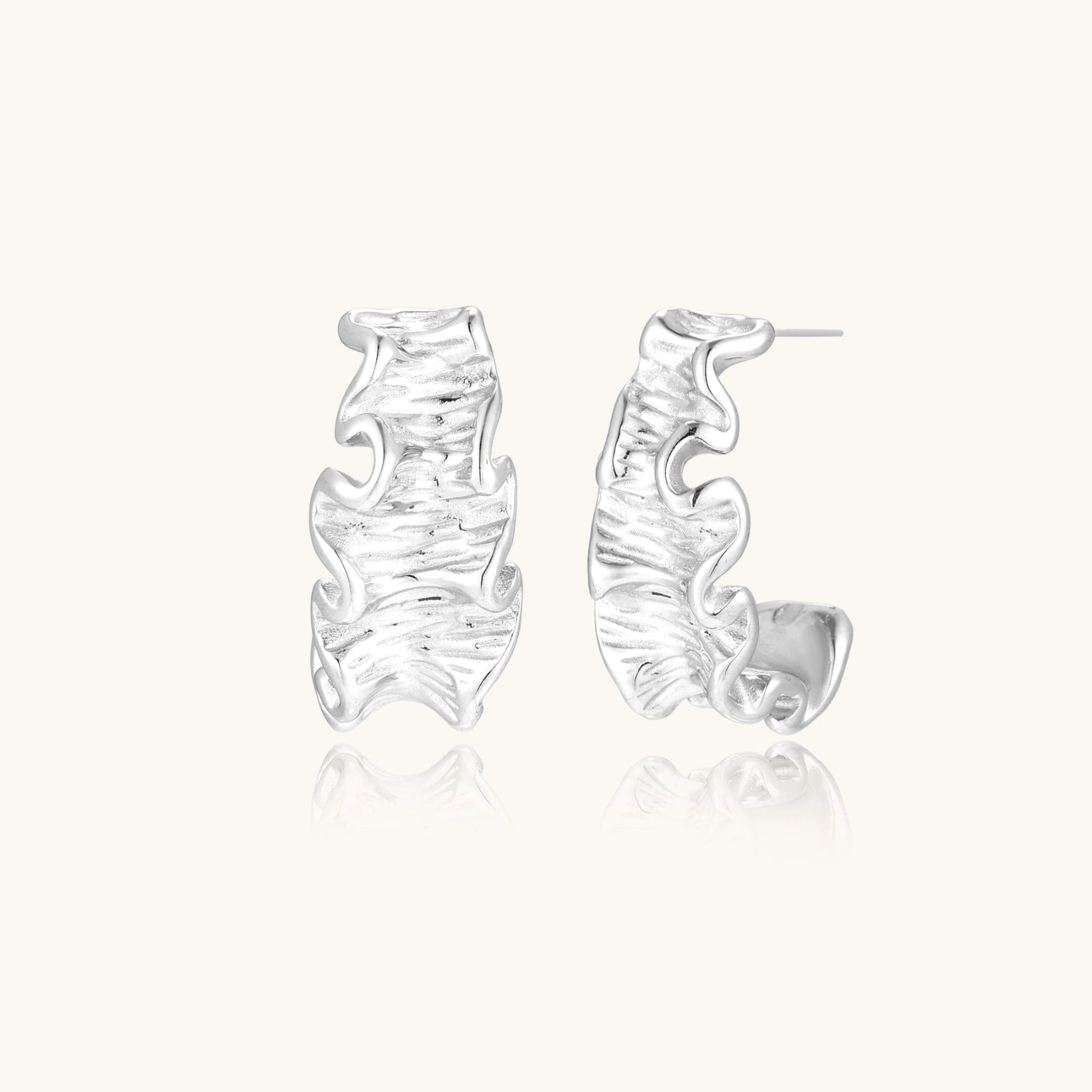 Textured white gold-plated hoop earrings with flowing wave pattern against cream background, showing dimensional rippled surface.