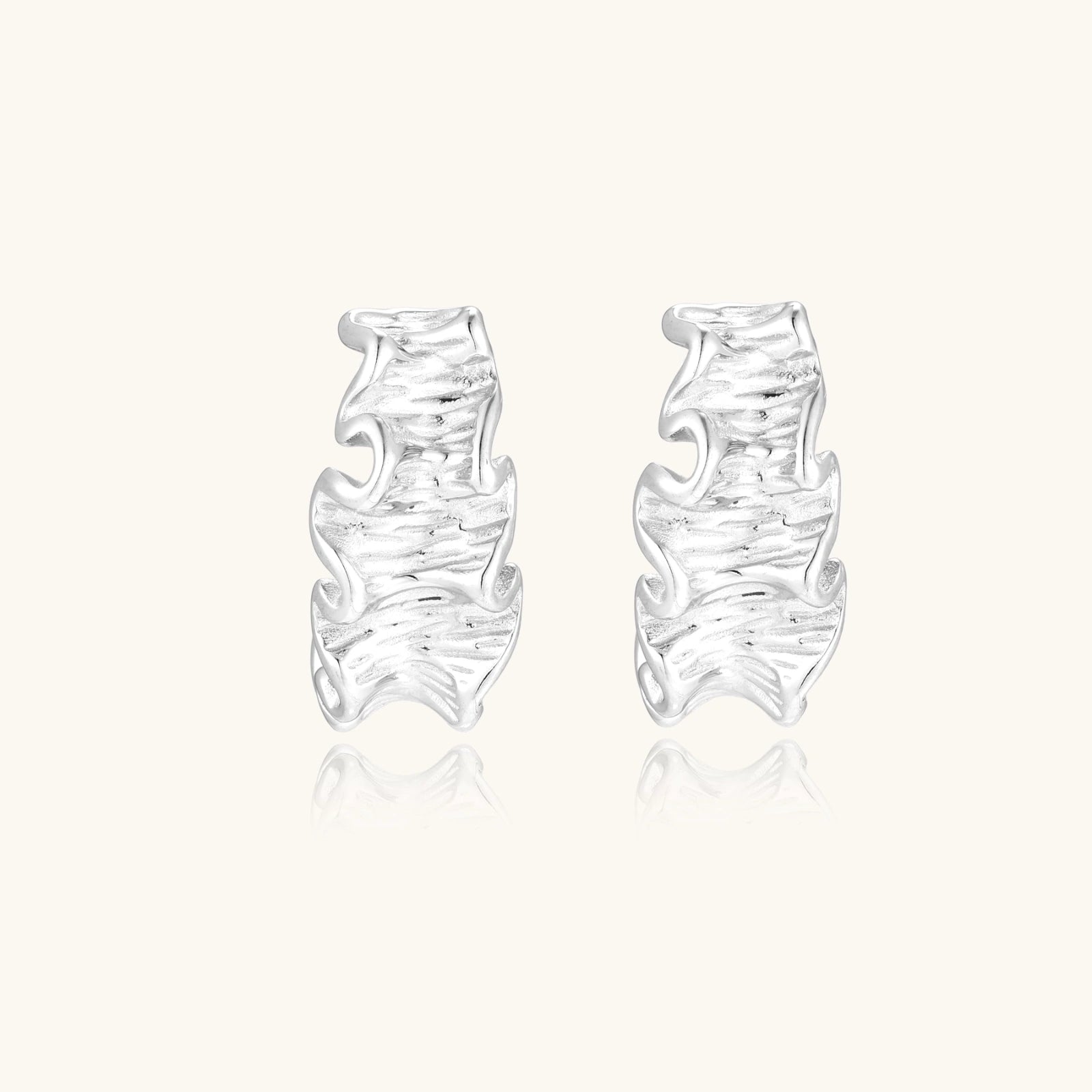 Textured white gold-plated hoop earrings with flowing wave pattern against cream background, showing dimensional rippled surface.