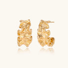 Pair of gold huggie hoops with sculpted wave texture and dimensional ripples on neutral background.