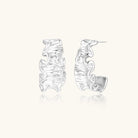 Textured white gold-plated hoop earrings with flowing wave pattern against cream background, showing dimensional rippled surface.