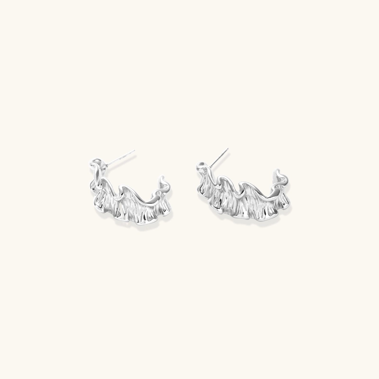 Textured white gold-plated hoop earrings with flowing wave pattern against cream background, showing dimensional rippled surface.