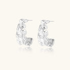 Textured white gold-plated hoop earrings with flowing wave pattern against cream background, showing dimensional rippled surface.