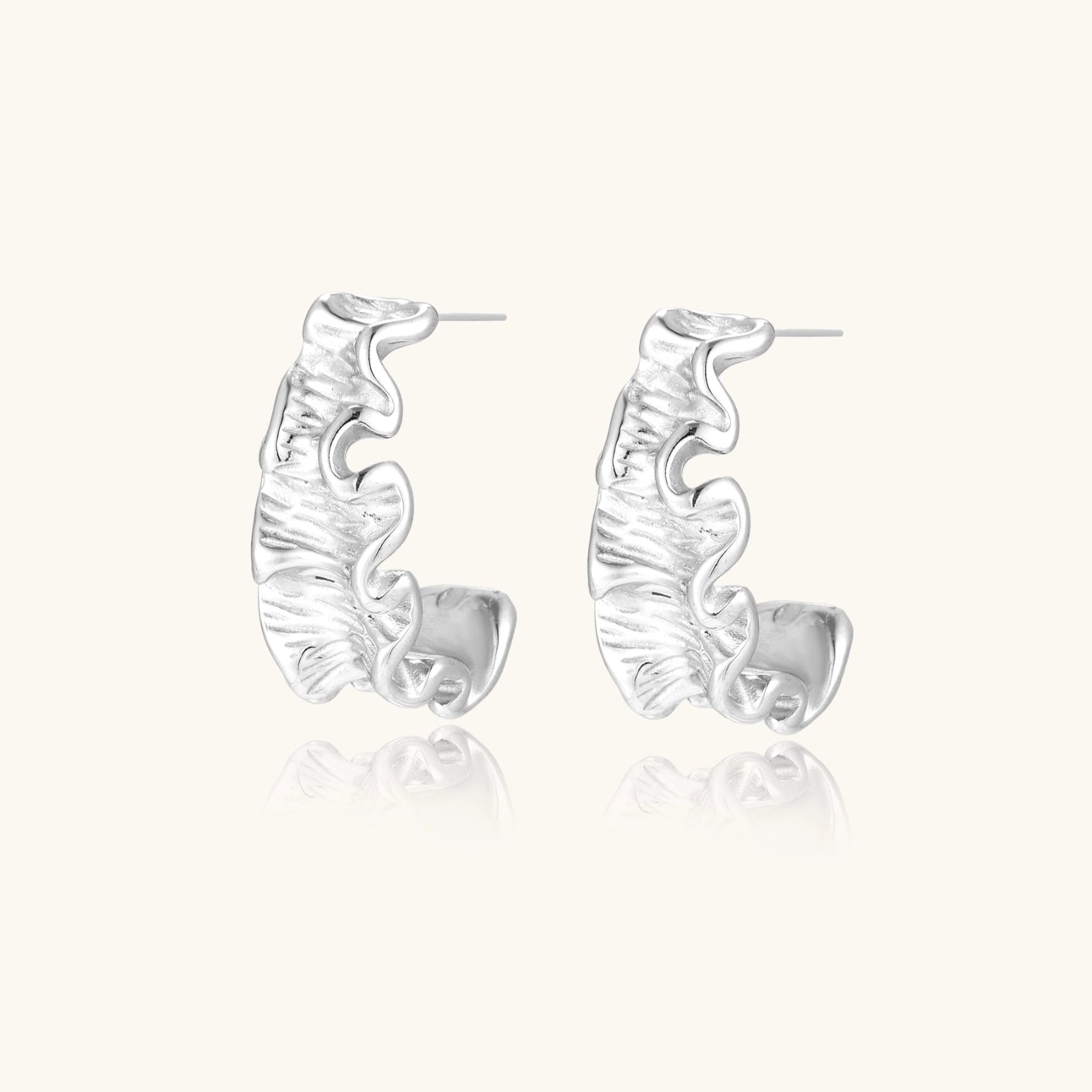 Textured white gold-plated hoop earrings with flowing wave pattern against cream background, showing dimensional rippled surface.
